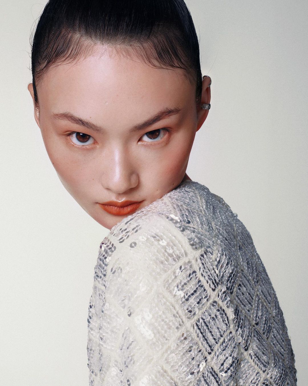 Close-up portrait of an Asian woman wearing a sequined jacket against a light-colored background, with focus on her neutral facial expression.