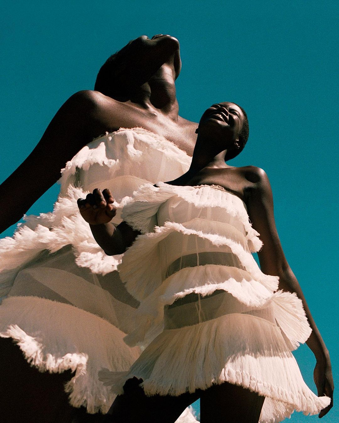 Two African women in white dresses, one looking up, smiling against a clear blue sky, captured with a low angle, creating a dreamy, elevated atmosphere.