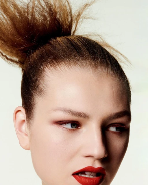 Close-up portrait of an adult female fashion model with red lipstick and styled brown hair against a pale green background in a beauty shot.