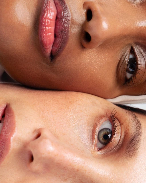 Close-up studio shot features two adult women with diverse skin tones, highlighting makeup, beauty, and skincare. Eye-level perspective.
