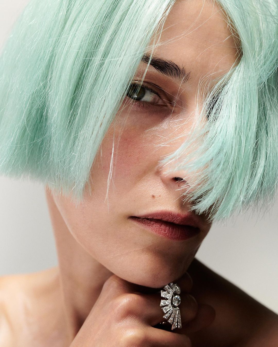 Close-up portrait of a young adult woman with short, mint-green hair wearing a statement diamond ring, set against a white background.