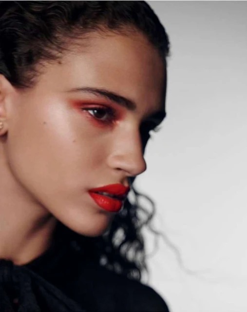 Close-up portrait of a young woman with bold red makeup on her lips and eyes, dark curly hair, and wearing a black top against a white background.