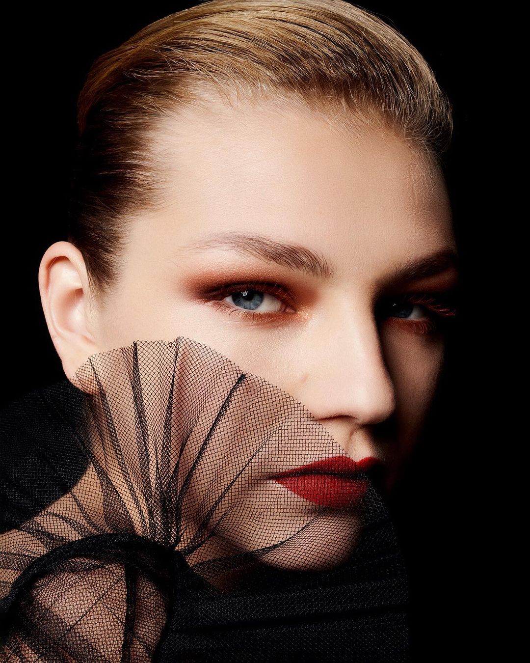 Close-up portrait of a woman with red lipstick and dramatic eye makeup partially obscured by black netting against a dark backdrop.