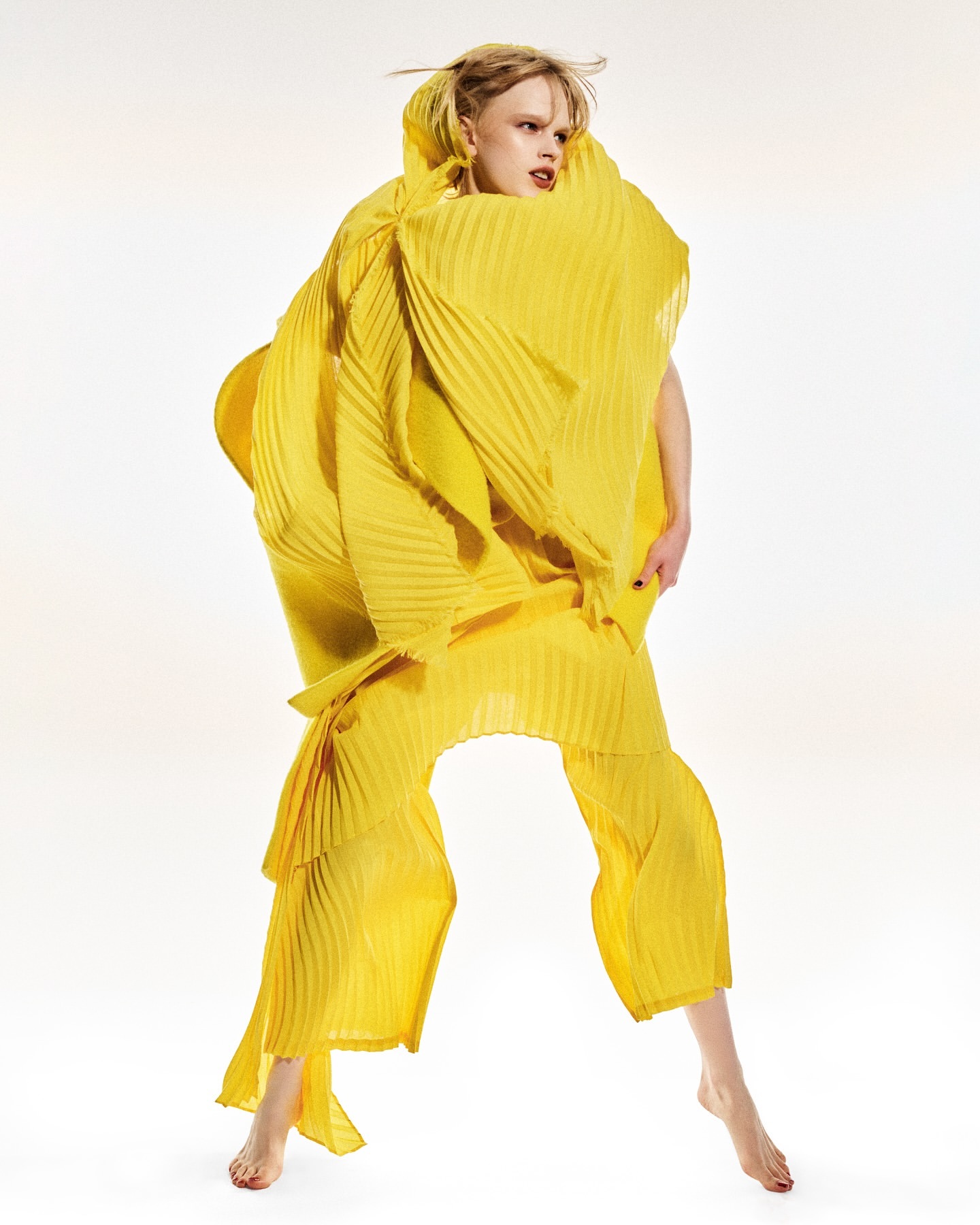 Fashion model in a bright yellow pleated dress with an oversized collar poses in a photo studio against a plain white background.