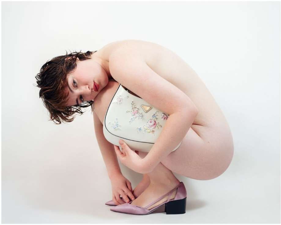 Young woman crouches holding white floral handbag with pink heeled shoes in a photo studio against a white background, artistic shot.