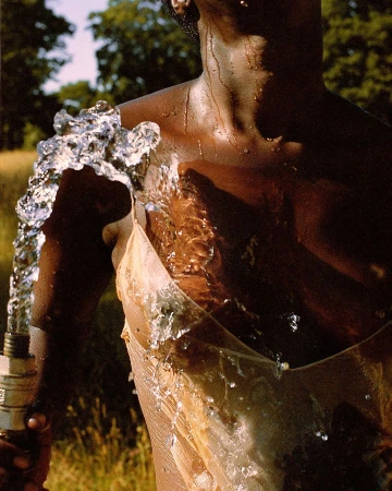 Close-up of a person with wet skin wearing a sheer slip dress, with a forceful arc of water splashing over their shoulder from a hose.