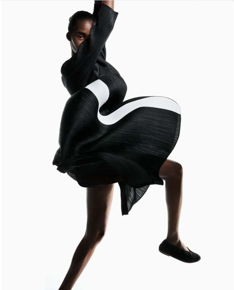 Fashion editorial shot of a Black model in a dynamic pose, wearing a black and white dress against a bright, clean white backdrop.