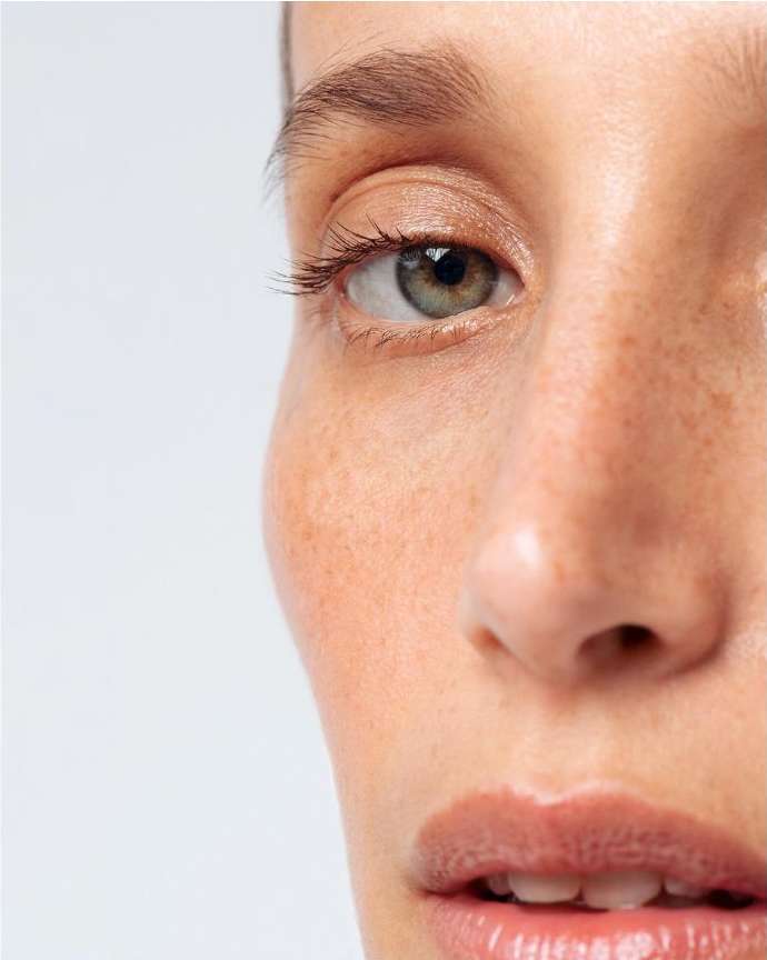 Close-up portrait of a woman with fair skin, visible freckles, and hazel eyes against a light background, showcasing beauty and skincare.