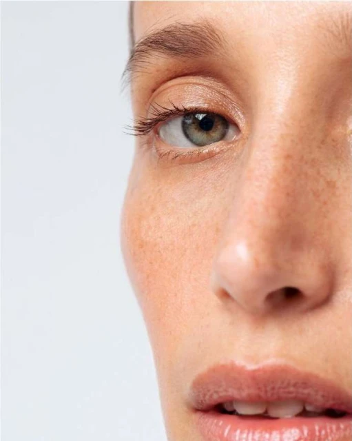 Close-up portrait of a woman with fair skin, visible freckles, and hazel eyes against a light background, showcasing beauty and skincare.
