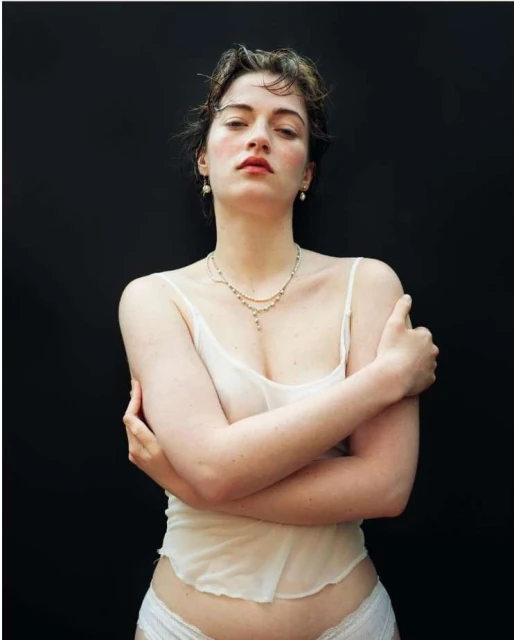 Portrait of a young adult woman in a sheer white top with her arms crossed, against a plain black background in a studio setting.