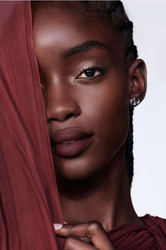 Close-up portrait of a Black woman holding a sheer burgundy fabric across the left side of her face, by Betina Du Toit.