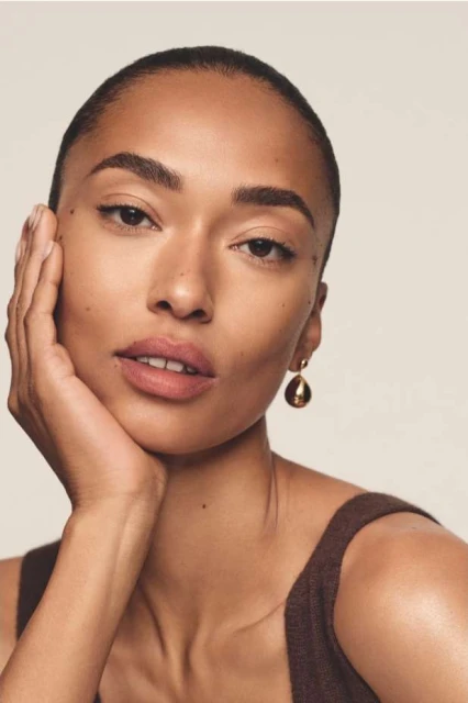 Close-up portrait of a woman with short hair and brown top looking at the camera, wearing gold earrings, and posing with her hand on her face.
