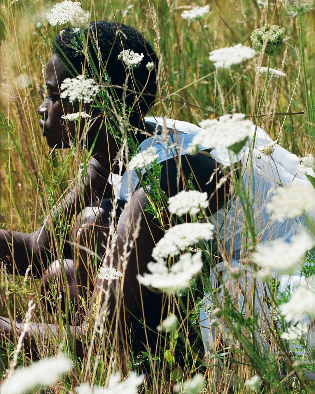 Portrait of a young black woman amid tall grass and white wildflowers on a sunny day. She wears a flowing white garment.