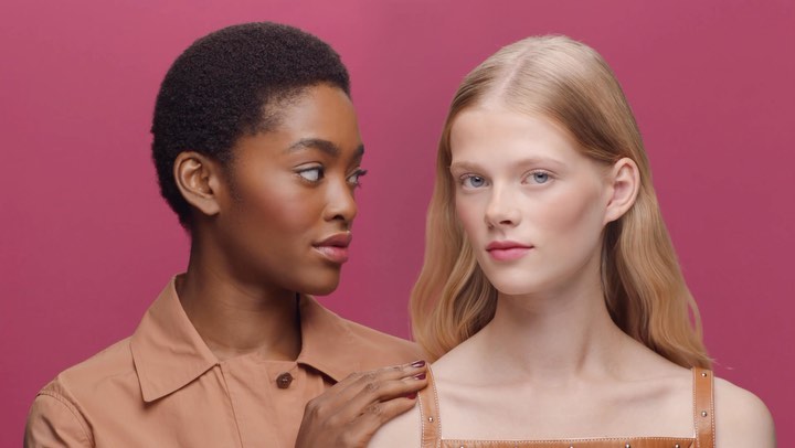 Two women pose in a studio shot. One woman, with dark skin and short hair, places her hand on the other woman's shoulder.