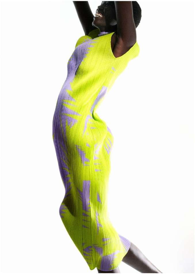 An adult woman in a sleeveless patterned midi dress smiles and raises her arms against a bright white background in a fashion portrait.