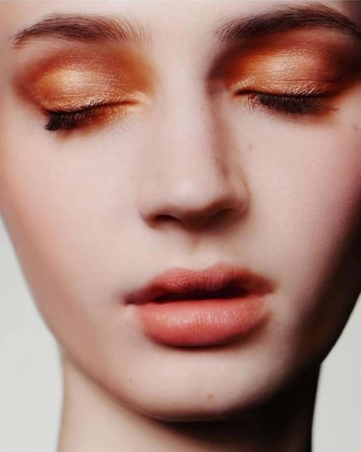 Close-up portrait of a young adult woman with closed eyes, shimmering copper eyeshadow, and natural lip color against a neutral background.