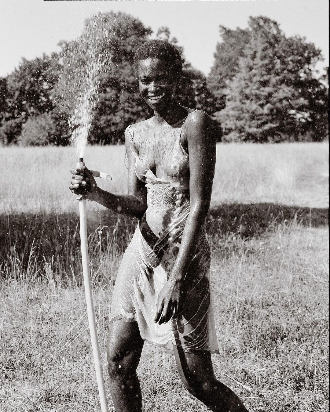 Woman stands smiling in a slip dress, holding a garden hose while spraying water on herself in an outdoor grassy area with trees.