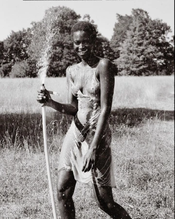 Woman stands smiling in a slip dress, holding a garden hose while spraying water on herself in an outdoor grassy area with trees.