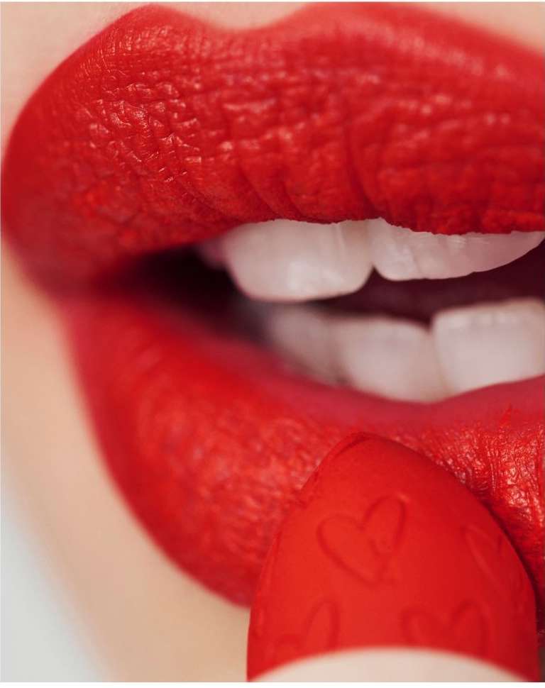 A close-up shot of a woman applying bright red lipstick, highlighting her teeth and the lipstick tube adorned with heart patterns.