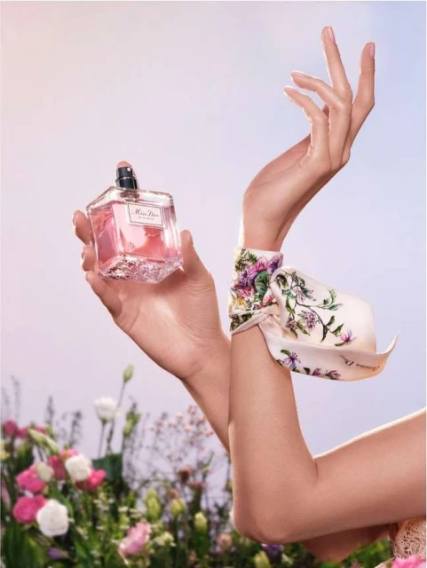 Close-up of a woman's hands holding a pink perfume bottle, wearing a floral scarf with flowers in the background on a light background.