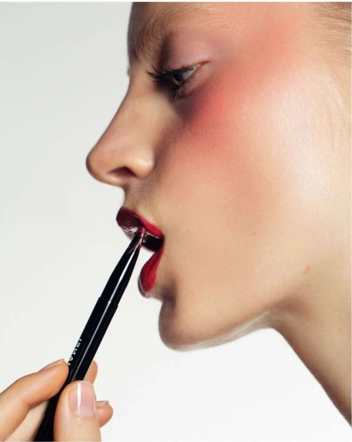 Woman with red lipstick holds tweezers to apply dark eye shadow for beauty makeup shoot, indoor studio setting and bright, even lighting.