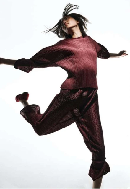 Fashion shot of a woman in a maroon plissé top and pants, jumping mid-air in a white studio. Movement and style emphasized.