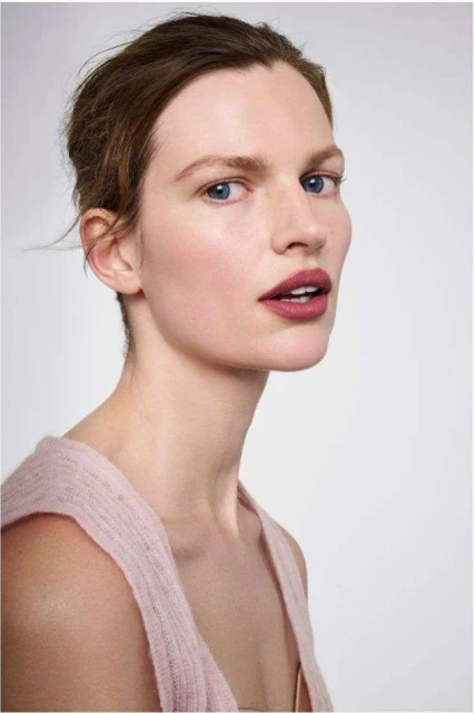 Close-up portrait of a young adult woman with fair skin, blue eyes, and light brown hair posing against a light gray background.