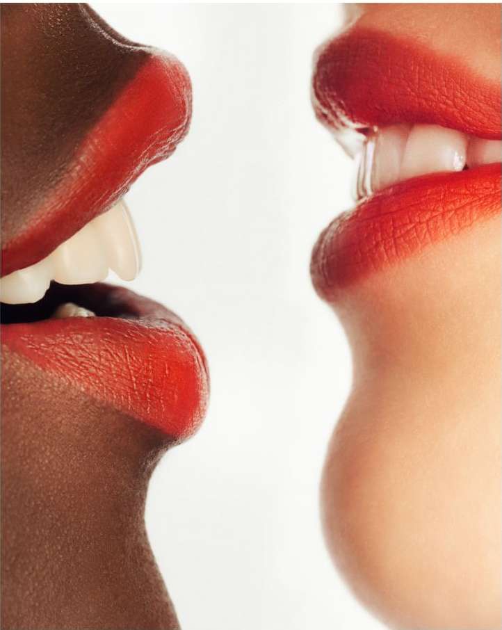 Close-up of two women of different ethnicities with red lipstick, teeth showing, against a white background, conveying a sense of diversity.