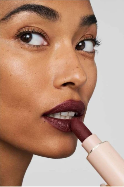 Adult woman with dark skin applies deep red lipstick close up against a neutral background in this studio beauty shot.