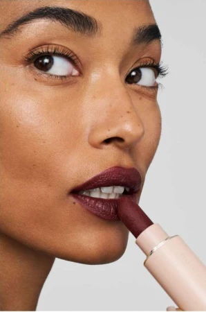Adult woman with dark skin applies deep red lipstick close up against a neutral background in this studio beauty shot.