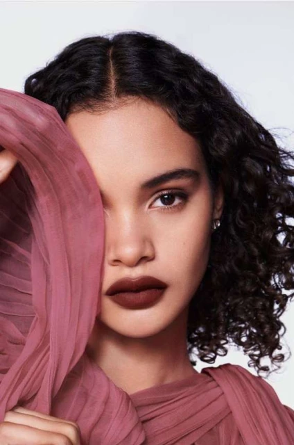 Close-up portrait of a young woman with dark curly hair and wearing dark lipstick, partially obscured by a draped pink textile.