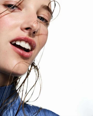 Close-up of a young woman with an open mouth and strands of hair on her face against a white background, wearing a blue top.