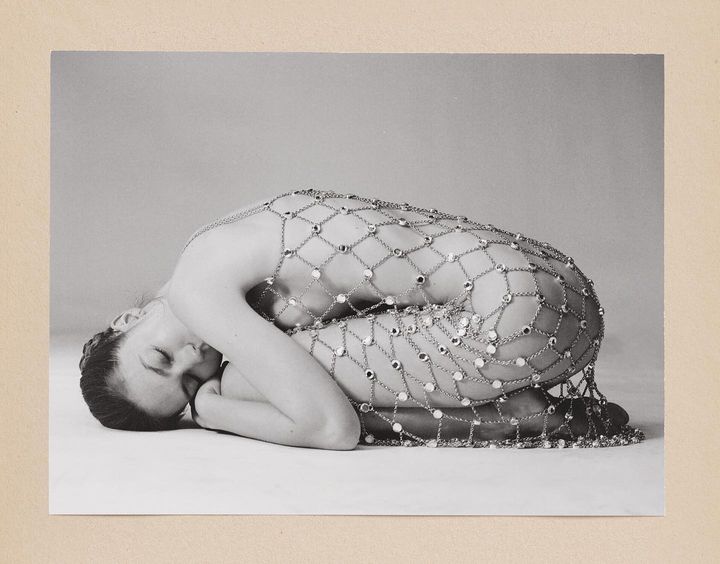 Monochrome portrait of a reclined woman covered in a mesh chain with jewels against a neutral background in a studio setting.