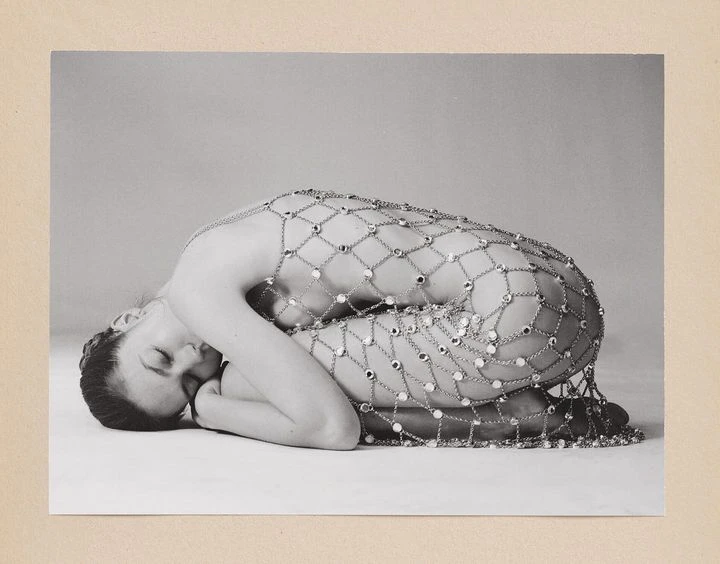 Monochrome portrait of a reclined woman covered in a mesh chain with jewels against a neutral background in a studio setting.