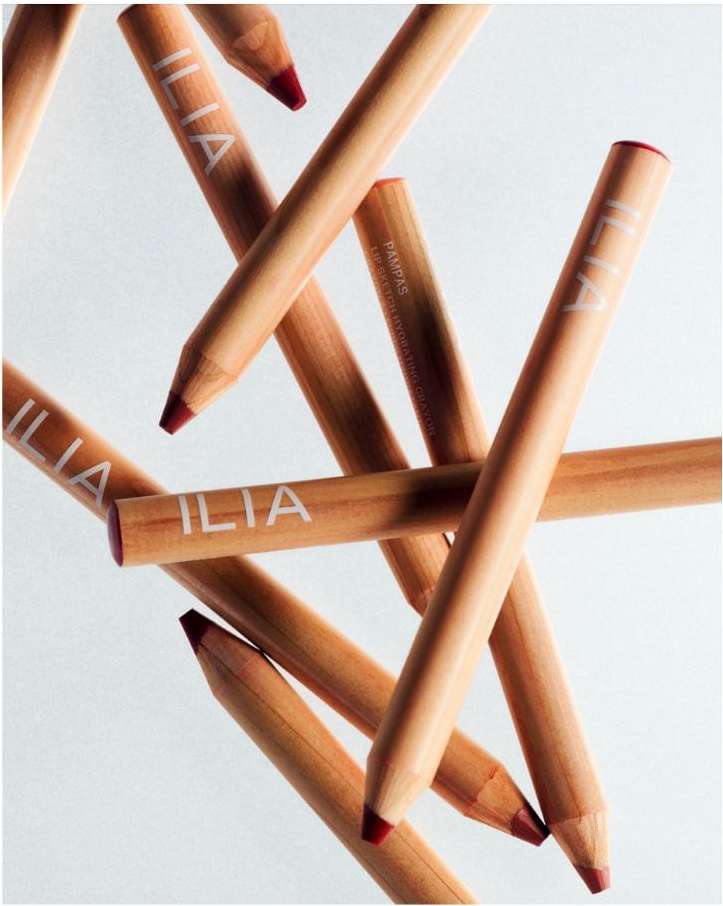 Close-up of 'ILIA' branded wooden lip crayons in a criss-cross pattern on a white background, red color at tips.