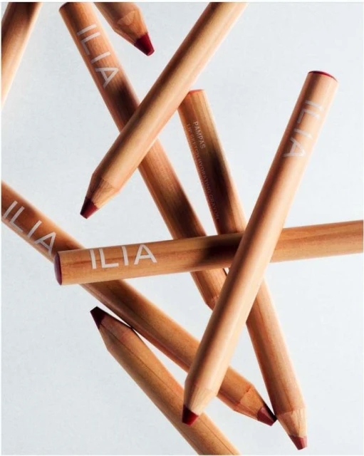 Close-up of 'ILIA' branded wooden lip crayons in a criss-cross pattern on a white background, red color at tips.