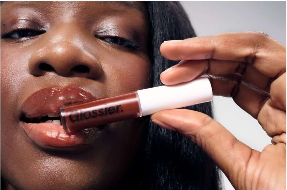 Close-up of a woman's face holding a Glossier lip gloss tube in front of her glossy lips, showcasing a beauty product indoors.