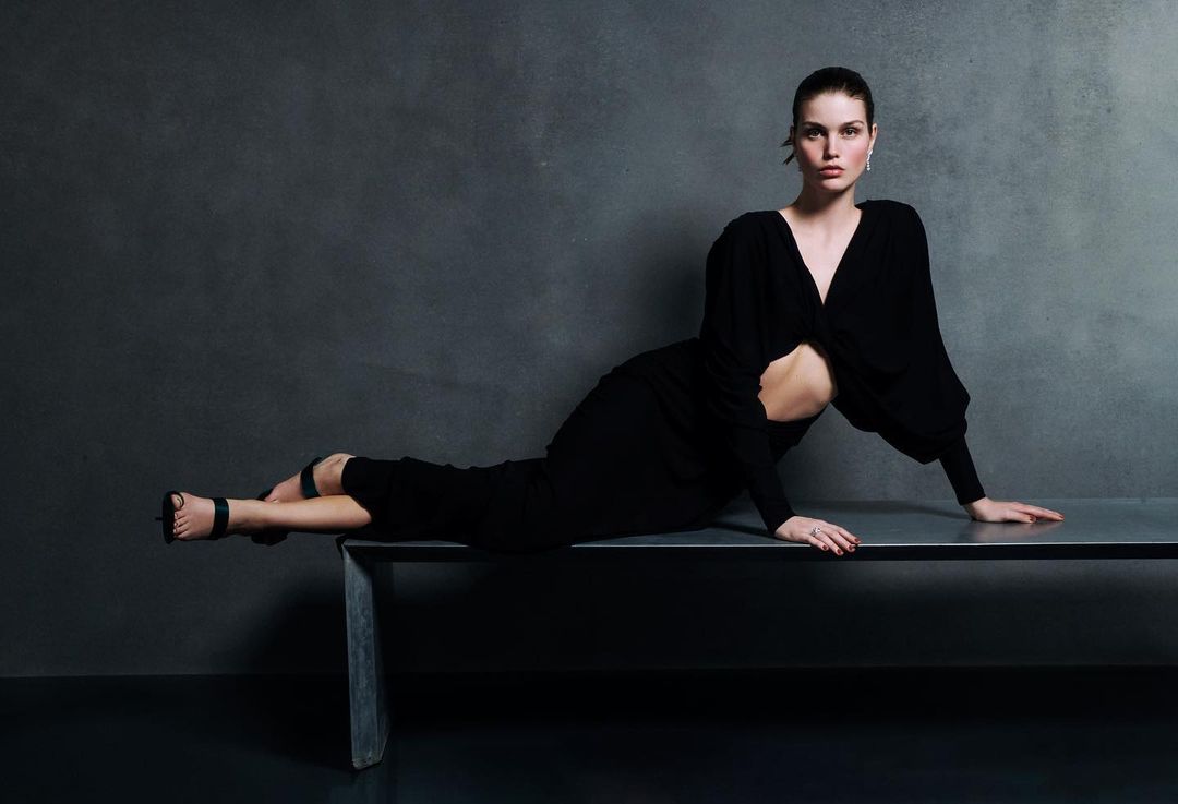 Woman in black dress reclines on a metal bench against a dark grey backdrop in a studio setting, showcasing fashion and style.