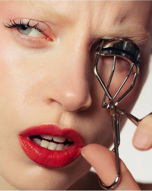 Close-up of a young woman applying mascara with a lash curler to her eye. She has red lipstick and fair skin, and she looks into the camera.