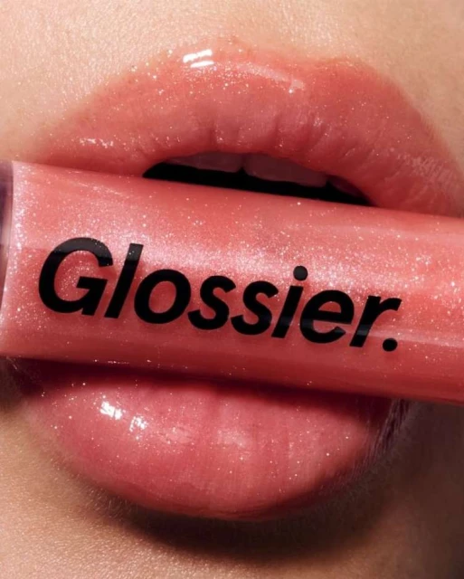 Close-up studio shot of an adult woman’s lips covered in glossy, sparkling lip gloss, with a tube of Glossier lip gloss obscuring her mouth.