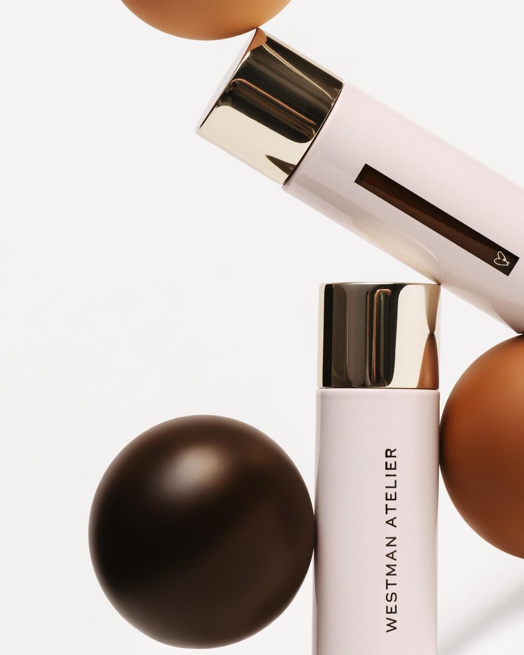 Still life shot of Westman Atelier makeup products with gold accents, alongside brown cosmetic spheres and a white background.