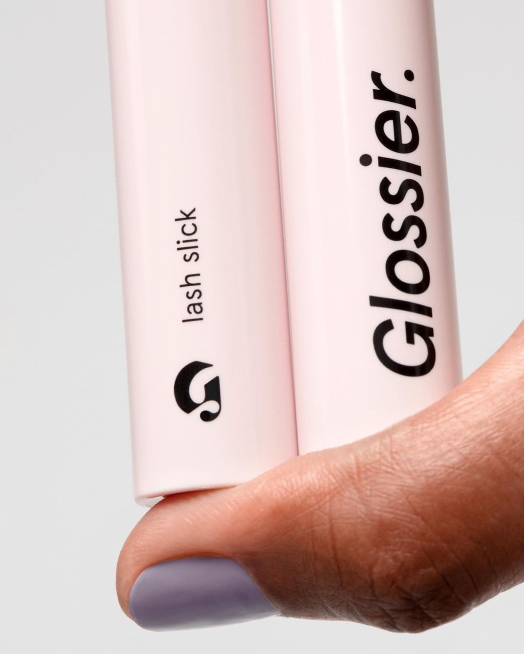 Close-up of two Glossier Lash Slick mascara tubes held by a hand, showcasing cosmetic beauty product details and branding.