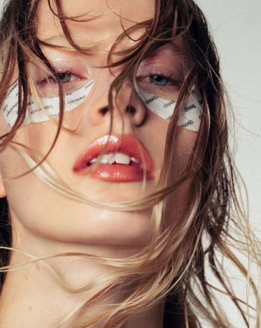 Close-up of a woman with blond hair, wearing glossy coral lipstick and eye patches printed with the brand name 'Chanel'. Beauty shot.
