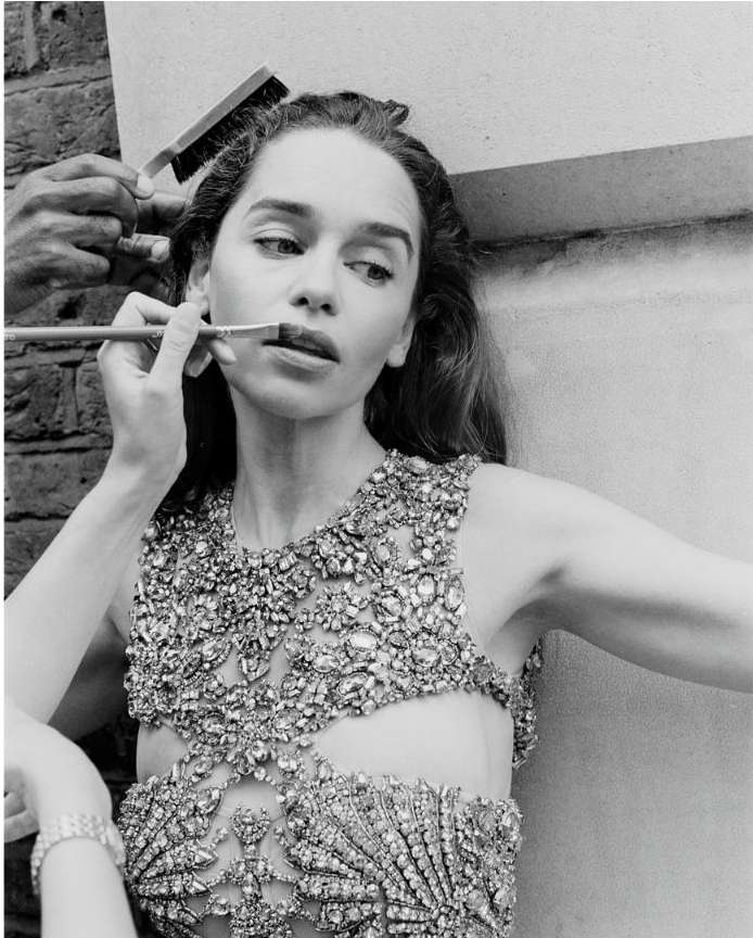 Black and white image of a woman having her makeup applied, featuring a jeweled top, makeup brush, and a person applying the makeup.
