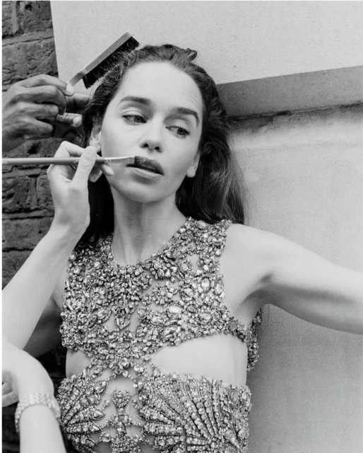 Black and white image of a woman having her makeup applied, featuring a jeweled top, makeup brush, and a person applying the makeup.