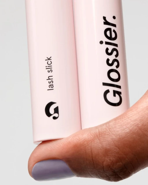 Close-up of Glossier Lash Slick tubes held by a person with light purple nail polish in a clean studio product shot.
