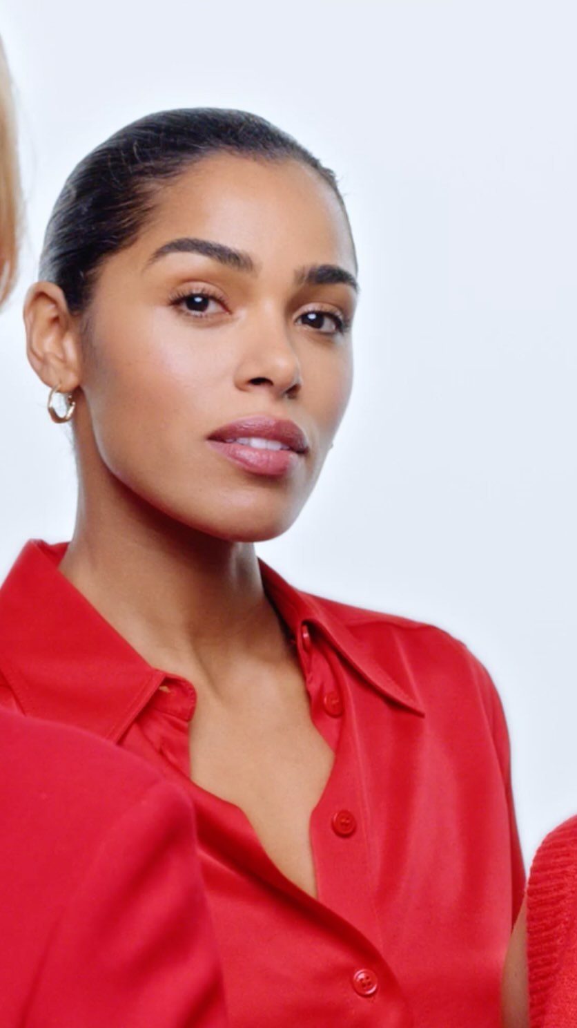 Portrait of a woman with dark hair and a red blouse in front of two other women against a plain white studio backdrop.