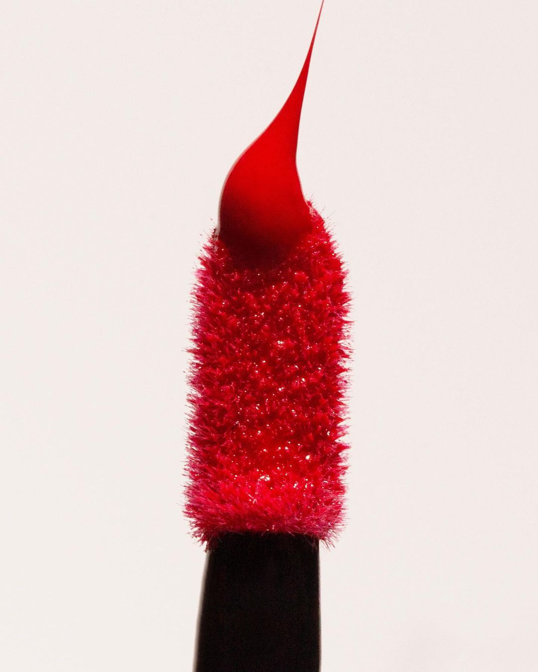 Close-up of a red lipstick applicator with a drop of product at the tip, isolated against a white background in a beauty shot.