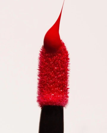 Close-up of a red lipstick applicator with a drop of product at the tip, isolated against a white background in a beauty shot.