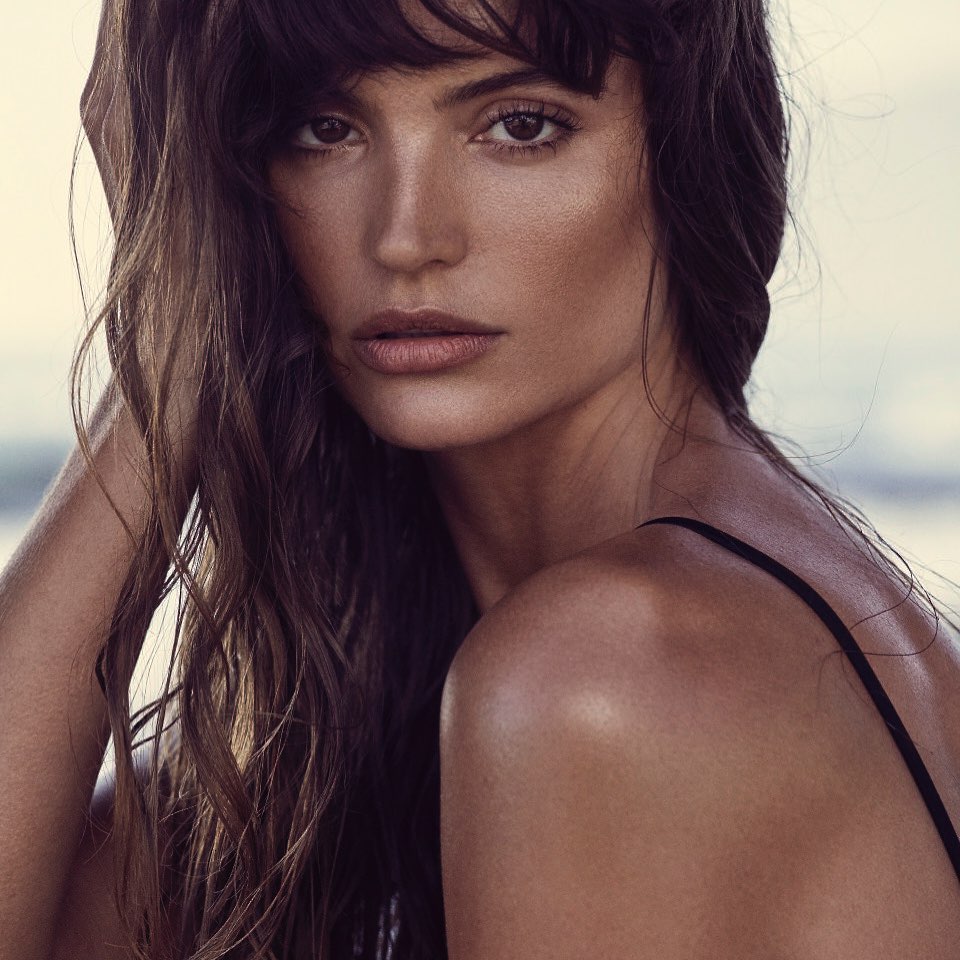 Close-up of a woman with long brown hair and bangs. She is looking at the camera with a neutral facial expression.